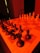 A chessboard is set up with black pieces on the left side and white pieces on the right, illuminated by a strong red light. The perspective is from one end of the board, giving prominence to the black pieces in the foreground. A knight and rook are clearly visible amongst the black pieces. The background is dimly lit with a shadowy chair in view.