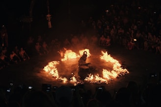 A person is seated in the center of a circle of fire, surrounded by a group of people. The spectacle takes place in a dimly lit environment with many onlookers gathered closely around, watching the performance. The flames create a dramatic and intense atmosphere.