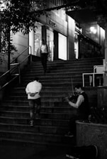 A violinist sitting at the bottom of a staircase, playing music. Two people are walking up the stairs, one looking at a phone and the other with hands behind his back. The scene takes place outside a store with large windows, and it appears to be evening.