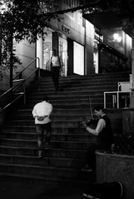 A violinist sitting at the bottom of a staircase, playing music. Two people are walking up the stairs, one looking at a phone and the other with hands behind his back. The scene takes place outside a store with large windows, and it appears to be evening.