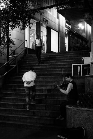 A violinist sitting at the bottom of a staircase, playing music. Two people are walking up the stairs, one looking at a phone and the other with hands behind his back. The scene takes place outside a store with large windows, and it appears to be evening.