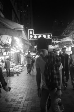 A black and white street scene depicting a busy night market with bright neon signs in an Asian language. Several people walk through the market, including a person in the foreground wearing a beanie and a plaid garment draped over one shoulder. Stalls line the sides of the street, displaying various goods.