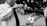 A young boy is playing a small trumpet-like instrument, while another child stands nearby looking up. The scene is set in a public area with several people in the background, including some sitting and standing. The image is in black and white, adding a timeless and candid feel.