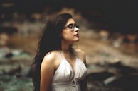 A person with long, dark hair and glasses stands in profile against a blurred natural background. They are wearing a white halter top and have a calm expression.