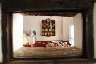 The interior of a rustic kitchen features a wooden shelf with neatly arranged plates and bowls, and a traditional oven in the foreground. A table is visible, covered with baskets of fruits and vegetables, evoking a sense of homeliness. Soft lighting enters through a partially opened window, illuminating the space.