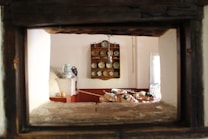 The interior of a rustic kitchen features a wooden shelf with neatly arranged plates and bowls, and a traditional oven in the foreground. A table is visible, covered with baskets of fruits and vegetables, evoking a sense of homeliness. Soft lighting enters through a partially opened window, illuminating the space.