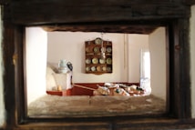 The interior of a rustic kitchen features a wooden shelf with neatly arranged plates and bowls, and a traditional oven in the foreground. A table is visible, covered with baskets of fruits and vegetables, evoking a sense of homeliness. Soft lighting enters through a partially opened window, illuminating the space.