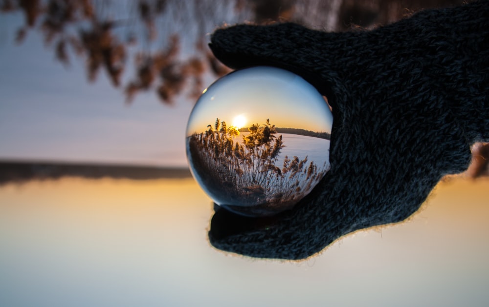 person wearing black gloves holding glass ball reflecting body of water with sunlight