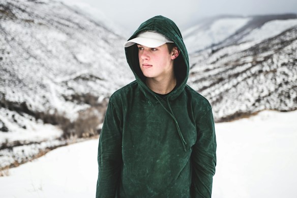 A person wearing a dark green hoodie and a white cap stands in a snowy mountainous landscape. The terrain is covered in snow with visible slopes and ridges in the background, creating a stark contrast to the dark clothing worn by the individual.