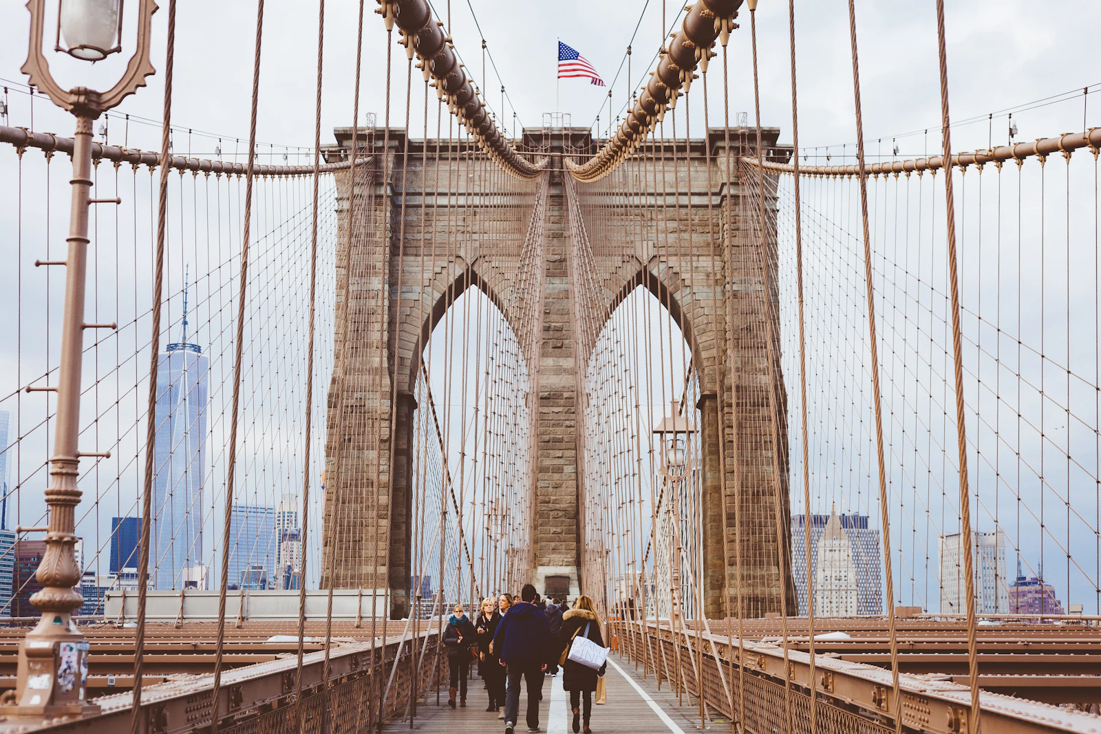 Travel photograph: Pedestrians walking towards a stone tower on the Brooklyn Bridge with lower Manhattan visible through the cables.