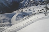 Snow-covered mountain slopes with skiing tracks, a small village in the background nestled among trees, and a metal tower structure on the right side. The landscape is marked by paths and fencing across the snowy terrain.