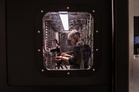 A person is standing inside a technical room or server room, working with equipment mounted on a rack. They appear focused and are adjusting or observing the setup. The equipment includes cables and a camera on a tripod. The scene is viewed through a square window with bolts, suggesting a secure or restricted area.