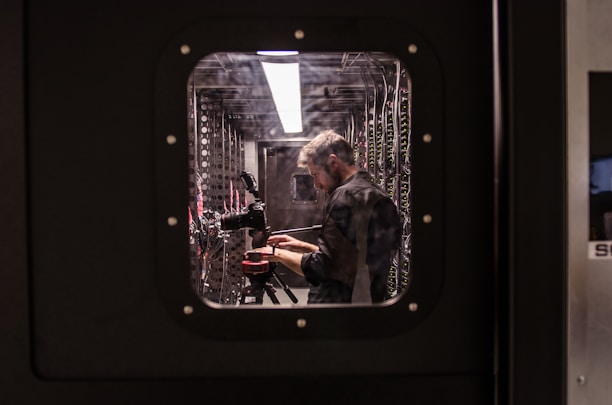A person is standing inside a technical room or server room, working with equipment mounted on a rack. They appear focused and are adjusting or observing the setup. The equipment includes cables and a camera on a tripod. The scene is viewed through a square window with bolts, suggesting a secure or restricted area.