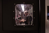 A person is standing inside a technical room or server room, working with equipment mounted on a rack. They appear focused and are adjusting or observing the setup. The equipment includes cables and a camera on a tripod. The scene is viewed through a square window with bolts, suggesting a secure or restricted area.