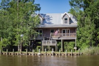 A wooden house is elevated on sturdy stilts, surrounded by lush green trees and vegetation. The house features a metal roof and an American flag hanging from the front porch. It is situated near a body of water, with a seawall and mooring posts visible in the foreground.