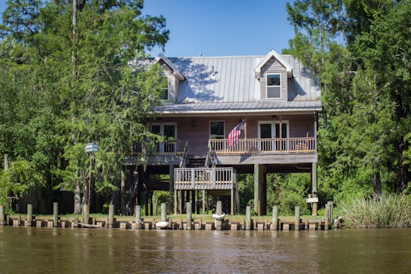 A wooden house is elevated on sturdy stilts, surrounded by lush green trees and vegetation. The house features a metal roof and an American flag hanging from the front porch. It is situated near a body of water, with a seawall and mooring posts visible in the foreground.