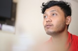 A man with short hair and a slight beard looks thoughtfully upward. The background is blurred, adding depth to the image. He is wearing a red shirt.