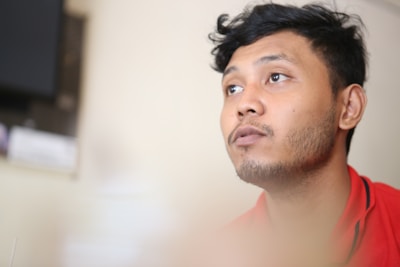 A man with short hair and a slight beard looks thoughtfully upward. The background is blurred, adding depth to the image. He is wearing a red shirt.