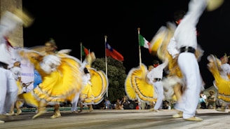 Dancers wearing vibrant yellow, white, and colorful traditional outfits perform energetically on stage. The movement of their skirts creates a dynamic blur effect. In the background, there are three flags of different countries, waving under the night sky.