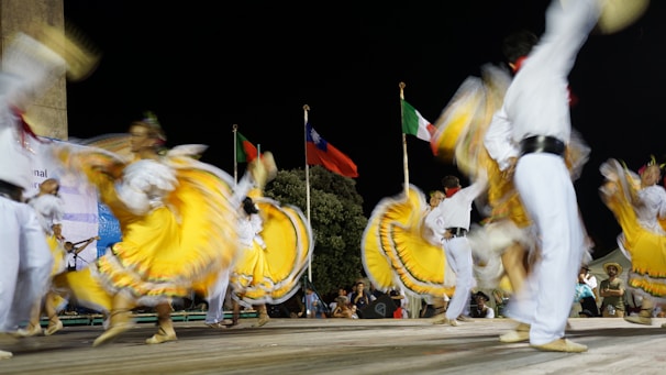 Dancers wearing vibrant yellow, white, and colorful traditional outfits perform energetically on stage. The movement of their skirts creates a dynamic blur effect. In the background, there are three flags of different countries, waving under the night sky.