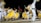Dancers wearing vibrant yellow, white, and colorful traditional outfits perform energetically on stage. The movement of their skirts creates a dynamic blur effect. In the background, there are three flags of different countries, waving under the night sky.