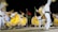 Dancers wearing vibrant yellow, white, and colorful traditional outfits perform energetically on stage. The movement of their skirts creates a dynamic blur effect. In the background, there are three flags of different countries, waving under the night sky.