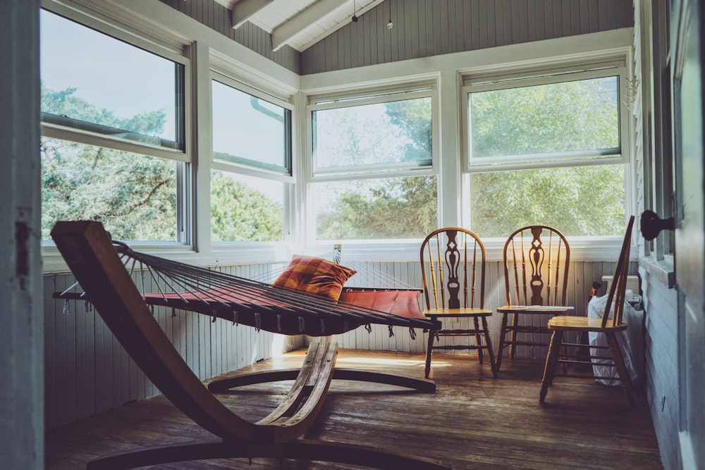 fotografia do interior da casa de madeira cinza, branca e marrom com três cadeiras Windsor de madeira marrom ao lado de rede marrom com janelas de vidro