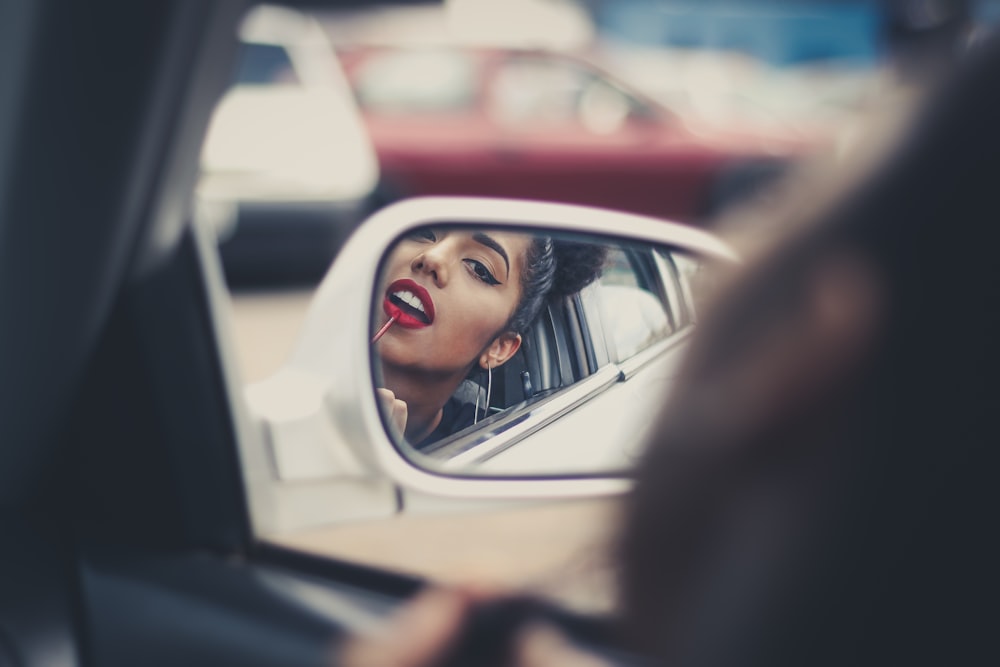 mulher colocando batom líquido em seus lábios enquanto olha para o espelho do veículo durante o dia