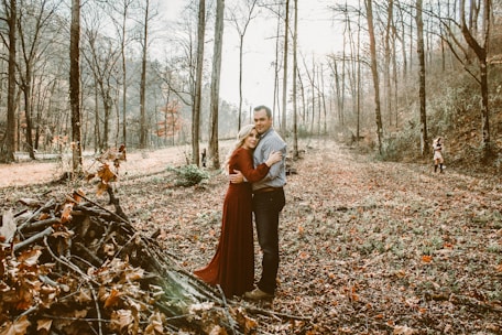 A couple embraces in a serene forest setting, surrounded by tall, bare trees and a carpet of fallen leaves. In the background, a child in a pink coat playfully runs. The scene is calm and portrays a close connection among the individuals.