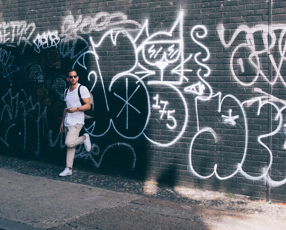 homme portant un t-shirt blanc à col rond appuyé sur un mur noir avec des graffitis blancs