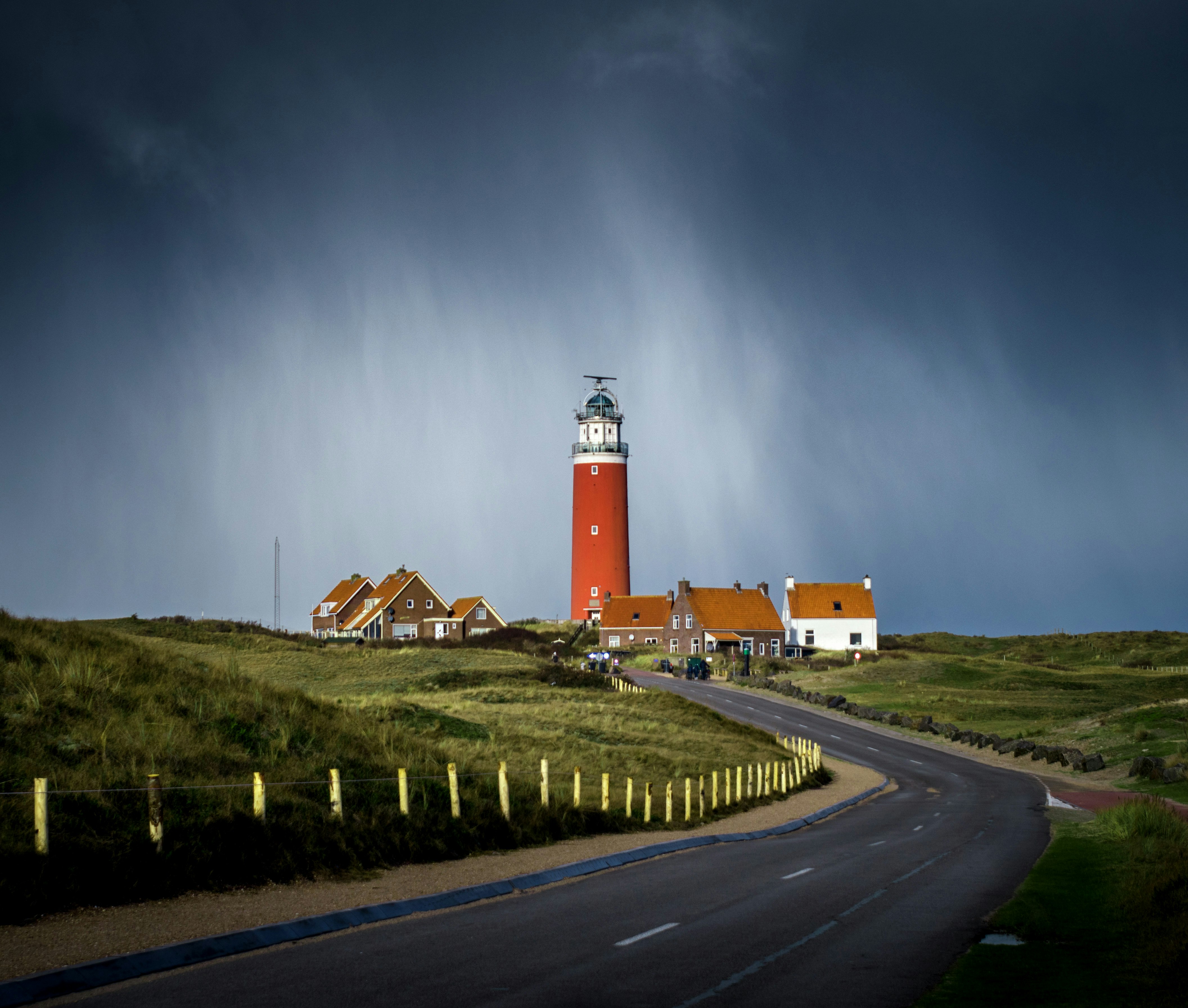 asphalt road between grass field going to brown lighthouse beside houses under cloudy sky