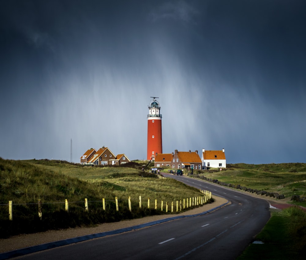 asphalt road between grass field going to brown lighthouse beside houses under cloudy sky