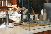A barista is preparing coffee using a pour-over method, surrounded by various coffee-making equipment on a marble countertop. Glass jars, a stack of paper coffee cups, and a coffee grinder are placed nearby, with a modern kitchen setup in the background.