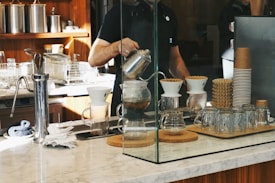 A barista is preparing coffee using a pour-over method, surrounded by various coffee-making equipment on a marble countertop. Glass jars, a stack of paper coffee cups, and a coffee grinder are placed nearby, with a modern kitchen setup in the background.