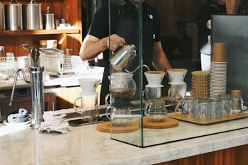 A barista is preparing coffee using a pour-over method, surrounded by various coffee-making equipment on a marble countertop. Glass jars, a stack of paper coffee cups, and a coffee grinder are placed nearby, with a modern kitchen setup in the background.
