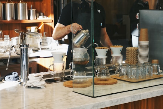 A barista is preparing coffee using a pour-over method, surrounded by various coffee-making equipment on a marble countertop. Glass jars, a stack of paper coffee cups, and a coffee grinder are placed nearby, with a modern kitchen setup in the background.