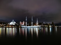 A well-lit sailboat is positioned on a calm body of water at night, creating a reflection in the water. Surrounding the boat are various illuminated buildings, including a church-like structure with a dome and another building with a tower. The sky is cloudy, casting a moody atmosphere over the scene.