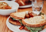 A sandwich with lettuce, tomato, and possibly bacon on whole grain bread sits on a white plate, secured with toothpicks. In the background, there is a bowl of mixed salad and a clear glass of water.