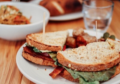 A sandwich with lettuce, tomato, and possibly bacon on whole grain bread sits on a white plate, secured with toothpicks. In the background, there is a bowl of mixed salad and a clear glass of water.