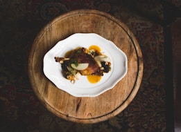 A beautifully plated dish featuring a roasted meat piece garnished with herbs and paired with vegetables and a vibrant sauce. The dish is presented on a white, decorative plate atop a rustic wooden table. The background consists of a patterned carpet, adding a touch of warmth and elegance to the setting.