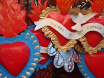 A collection of decorative flaming hearts, with intricate designs and bright colors. A white dove with spread wings is perched on one of the hearts, which is wrapped in a white ribbon bearing the text 'Vivir&aacute;s en mi coraz&oacute;n'. The hearts are adorned with blue, red, and gold colors, with some displaying flame-like projections at the top.