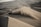 A person on a quad bike is riding up a large, sandy dune in a desert. The vehicle is kicking up a significant amount of dust and sand as it moves. The landscape is filled with high dunes and there is a sense of vastness and isolation.
