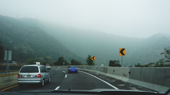 A winding road with several cars driving along it, surrounded by dense fog that obscures the view of the forested hills in the background. Bright yellow and black road signs indicate a curve ahead, suggesting careful driving due to limited visibility.