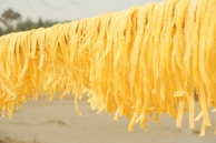 Long strands of yellow pasta are hanging to dry on a rod against a blurred outdoor background. The pasta appears fresh and handmade, with a vibrant color.