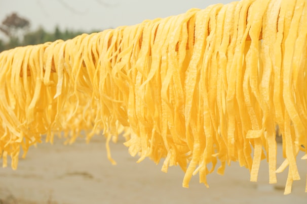 Long strands of yellow pasta are hanging to dry on a rod against a blurred outdoor background. The pasta appears fresh and handmade, with a vibrant color.