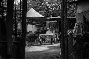 A rustic outdoor setting with a canopy overhead, a table covered with a patterned tablecloth, and plastic chairs scattered around. The area is enclosed by structure supports and netting, with plants visible on the right side. The background shows a modest building with corrugated metal roofing.