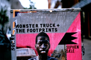 A weathered poster on a street cabinet featuring a person with a serious expression. The poster includes text promoting a music event, 'Monster Truck + The Footprints,' along with the venue name 'SOPHIEN S&AElig;LE.' The image possesses a gritty, urban feel with a mix of vibrant and muted colors.