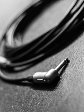 A close-up shot of a coiled audio cable with a quarter-inch jack resting on a textured surface. The cable is partially in focus with a black and white color scheme, highlighting the metallic and rubber parts of the plug.
