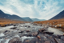 A serene mountain landscape featuring a flowing river with clear, glistening water running over smooth stones and pebbles. The river is surrounded by grassy banks with a backdrop of towering mountains under a cloudy sky. The scene captures the tranquility and natural beauty of a remote wilderness.