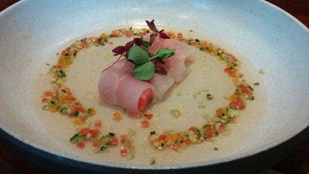 A dish featuring a piece of raw fish delicately rolled and topped with microgreens, surrounded by a colorful mixture of finely chopped vegetables. This is presented on a textured plate with a light sauce at the base.